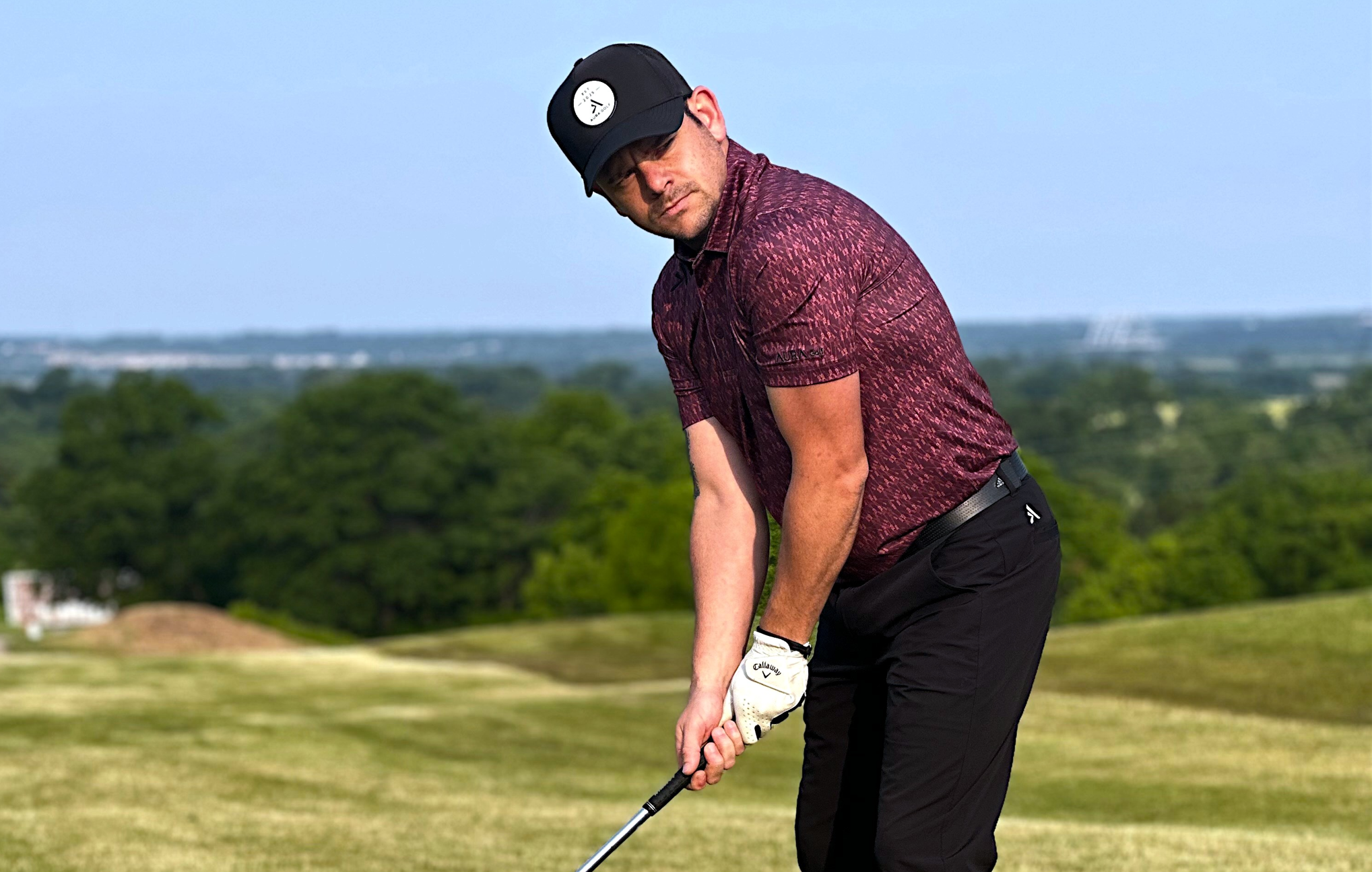 Golfer in a maroon shirt and black pants on a golf course with trees and sky in the background